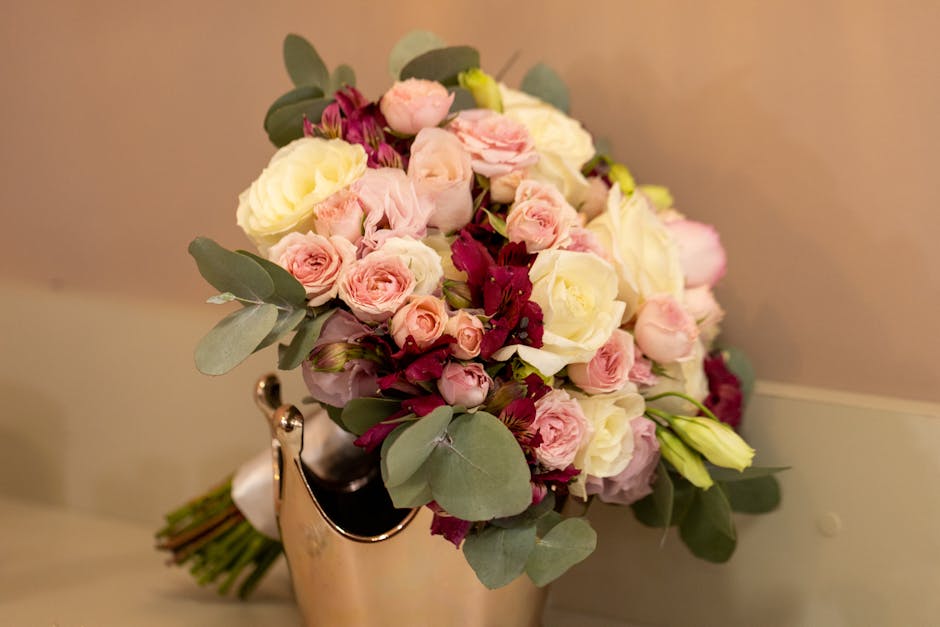 A bouquet arrangement featuring a mix of fresh pale pink roses, white roses, and small pink spray roses, accented with dark red flowers and lush eucalyptus leaves. The flowers are tightly packed, show