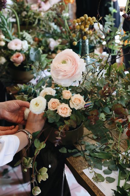 Close-up of a florist's hand adjusting a floral arrangement featuring pale pink and cream ranunculus flowers, complemented by sprigs of eucalyptus with silvery-green leaves. The arrangement is set on 