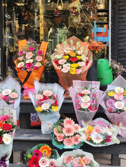 A variety of fresh flower bouquets displayed outside a flower shop window, featuring vibrant blooms such as pink, yellow, white, and red gerbera daisies, roses, and chrysanthemums. Each bouquet is wra