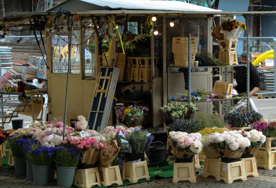 A street-side flower stall showcasing a vibrant arrangement of fresh flowers. The display includes pink and white peonies, purple delphiniums, and blue cornflowers, all wrapped in brown paper and plac