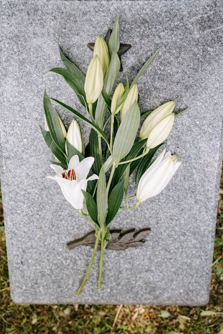 A floral tribute consisting of white lilies with open petals and prominent red stamens, and closed white lily buds, arranged on a light grey granite memorial stone. The flowers are positioned on a woo