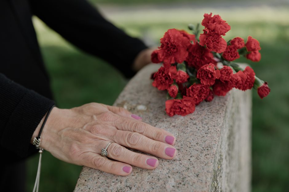 A close-up of a woman’s left hand resting on a stone ledge, displaying a silver ring with a gemstone, and nails painted in soft pink. Next to her hand, a small bouquet of bright red carnations with